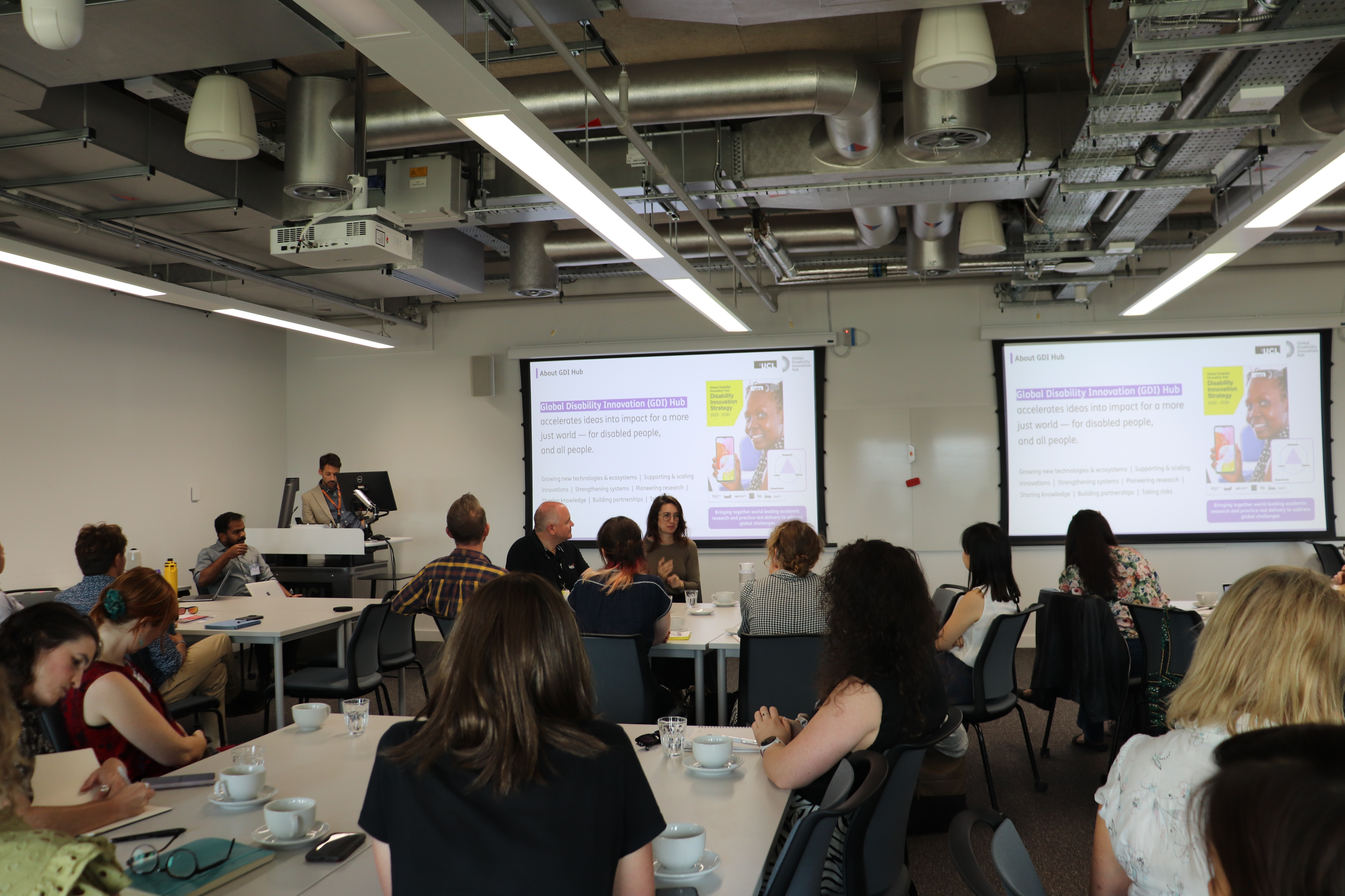 Iain McKinnon, Co-founder and CEO of GDI Hub presents about the Global Disability Innovation Hub. Participants are seated at tables, watching the presentation on a screen. A BSL interpreter signs as Iain speaks.  