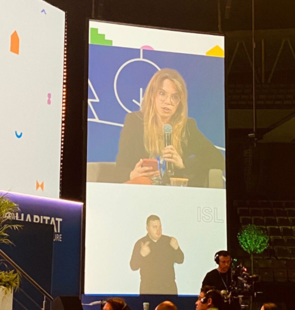 A large screen at the conference showing speaker Mikaela Patrick and International Sign Language interpreter