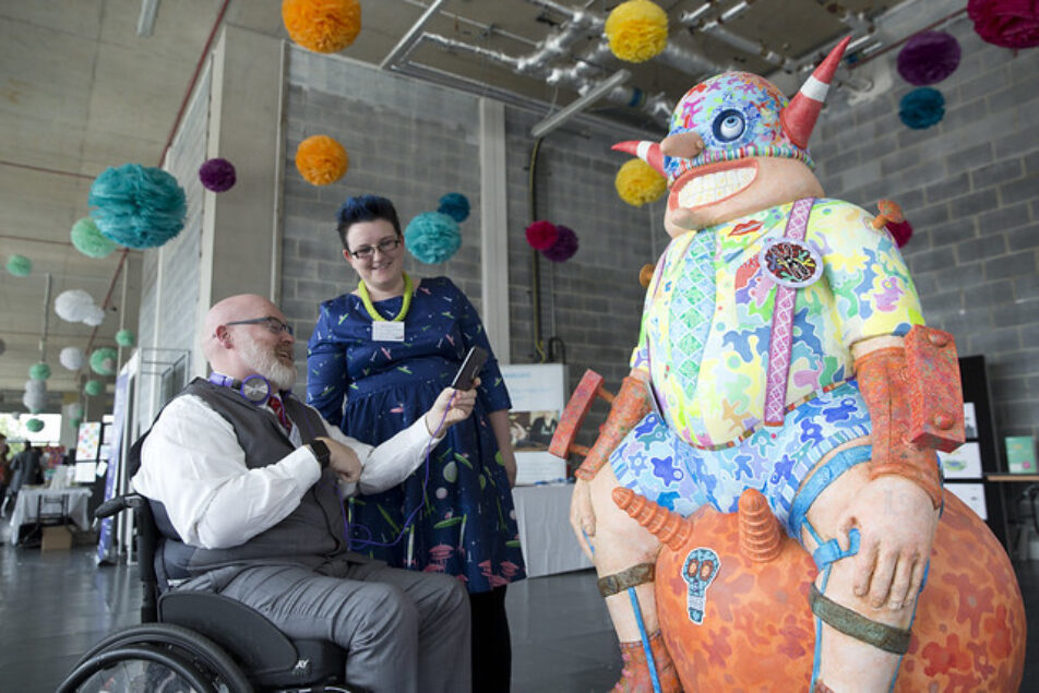 An old man in a wheelchair is taking a photo of an art sculpture of a man wearing shorts and braces on a space hopper.