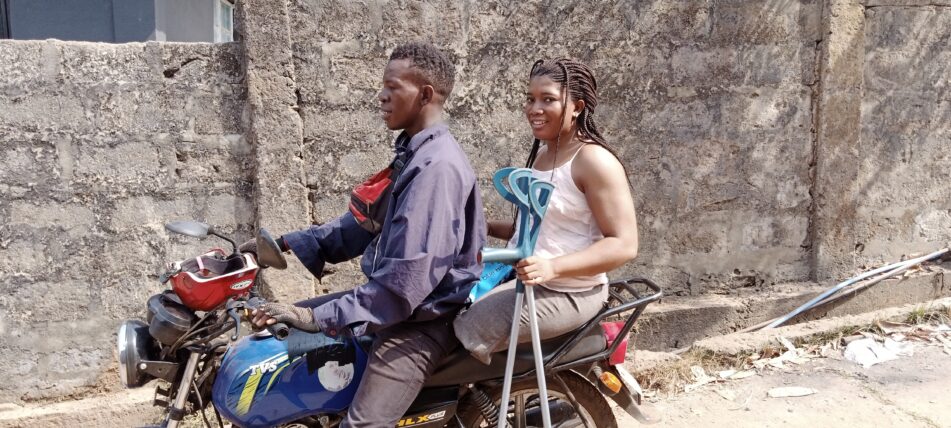 Image of two people on a motorbike in freetown. A man is driving, and a female passenger is holding walking aids.