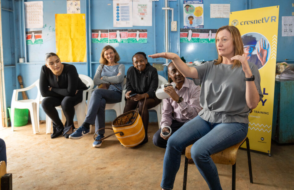 A woman in a grey top and jeans is speaking and gesturing with her hands while sitting on a chair. Four other people are seated behind her, looking at her. A yellow banner with "cresnetVR" is visible in the background.