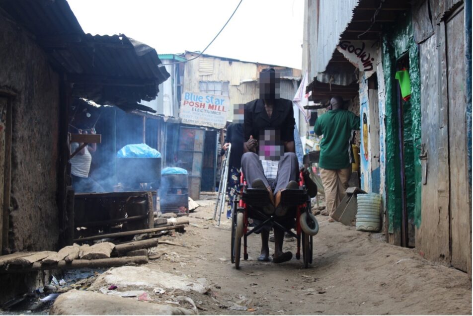 A person pushing the wheelchair with another person sitting on it on a  narrow alley in an informal settlement