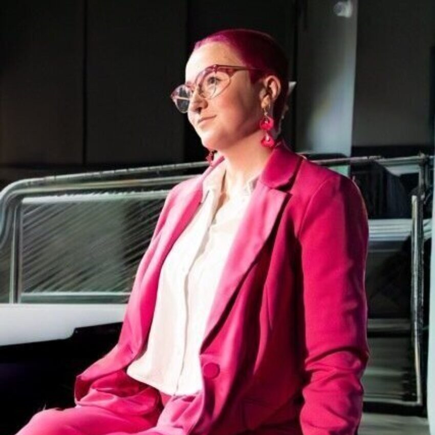 Jet in a bright pink suit sits in a wheelchair, wearing matching accessories and a white blouse, positioned in a modern indoor setting with metal railings in the background.