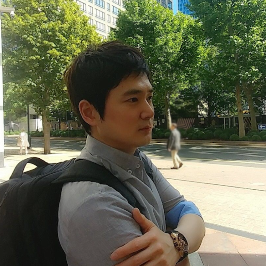 Dr Youngjun Cho looking away from the camera with arms folded. Trees and buildings in the background.