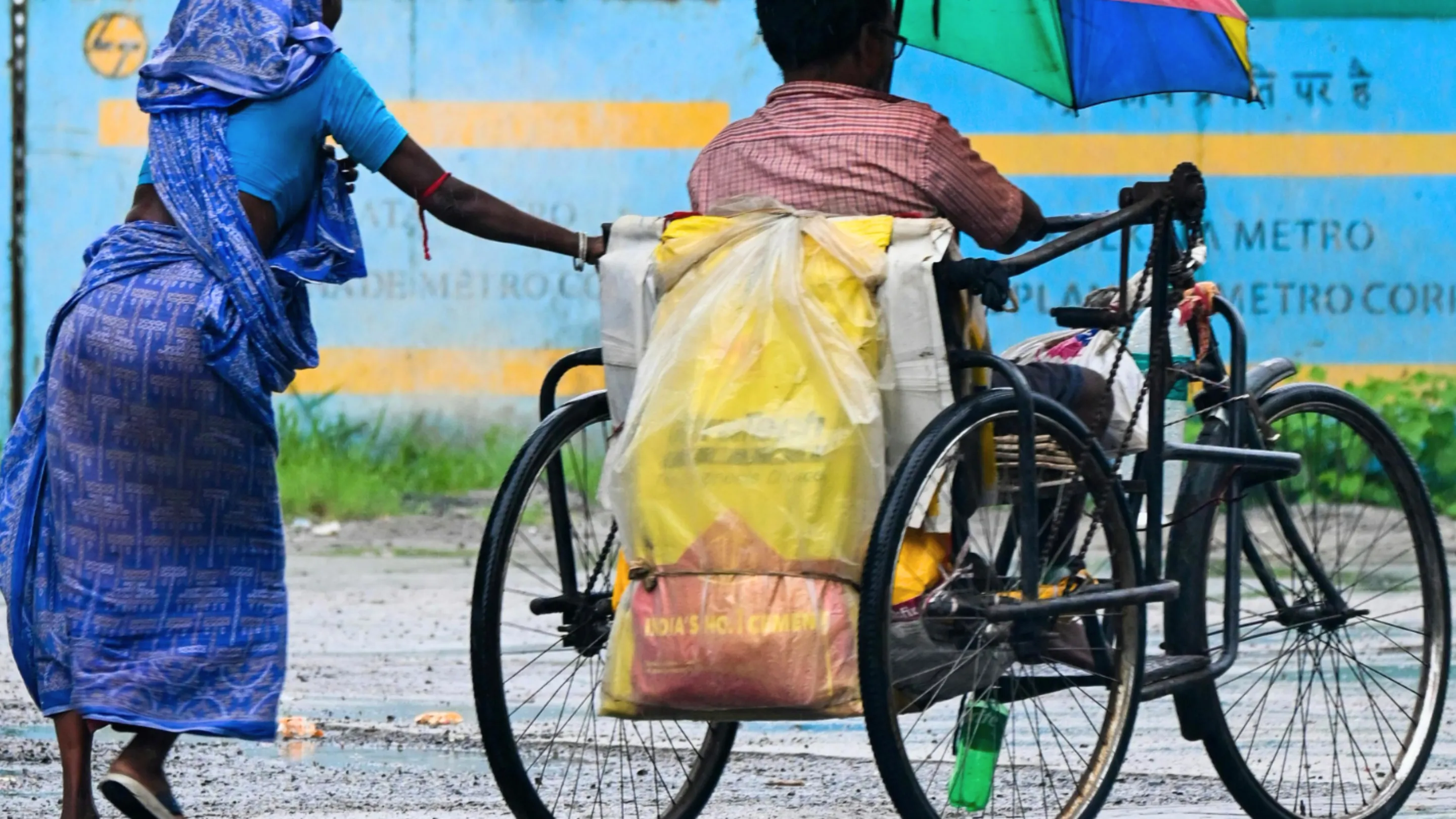 A person in a blue sari walks alongside a person on a home-built wheelchair, holding onto the back of the wheelchair. The person on the wheelchair holds a green and blue umbrella.