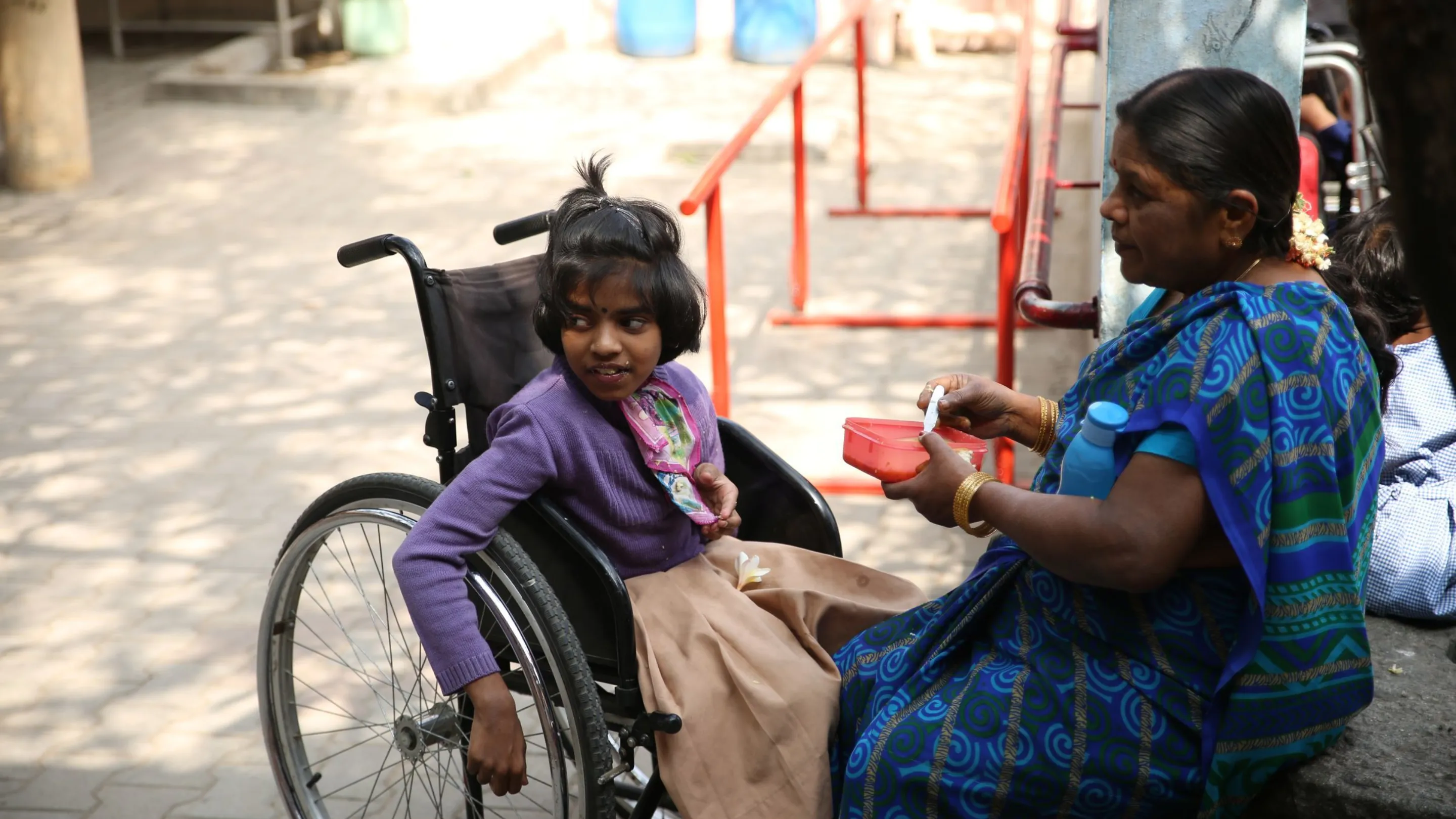 Photo of a young Indian girl on an oversized wheelchair being fed by her mother