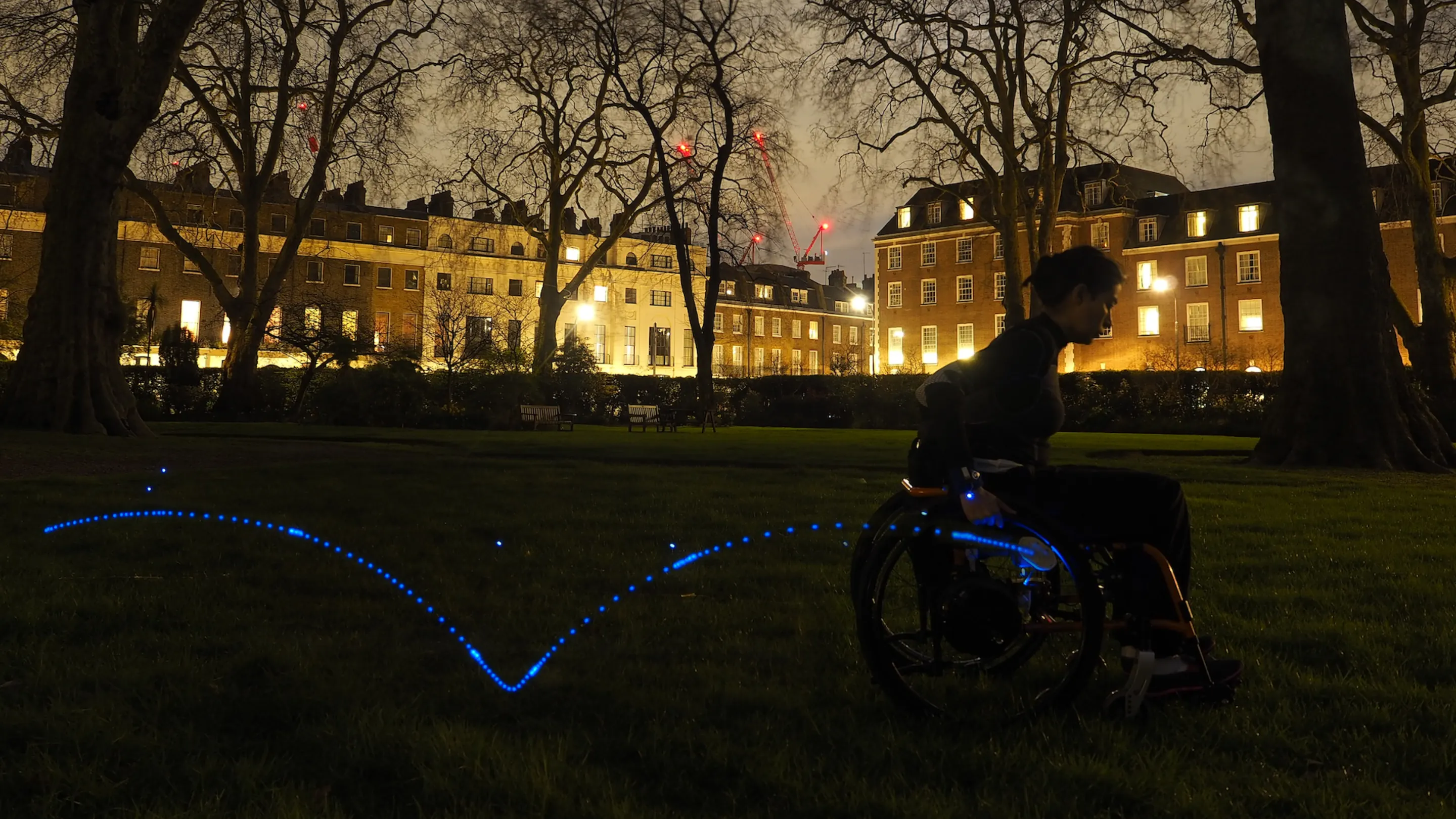 A view of a park in London at night. A manual wheelchair user is propelling a wheelchair in this park. The wheelchair user is wearing an inertial sensor in the wrist, a second one is attached to the wheel. A trail of blue light shows the trajectory that the sensor measures from a couple propulsions of the wheelchair.