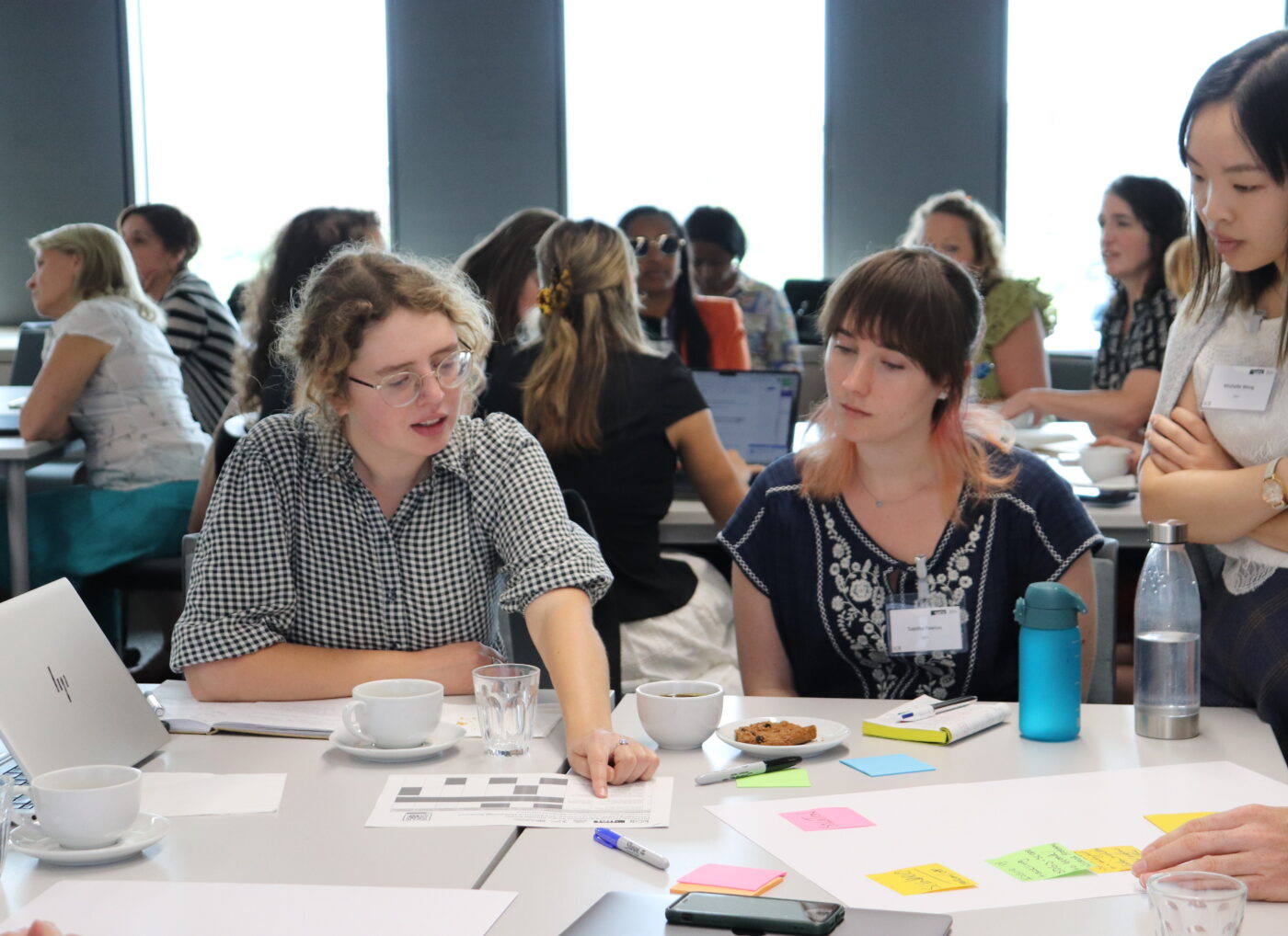 A diverse group of adults are seated at tables in a bright, modern room. Other people are visible in the background, some working on laptops.