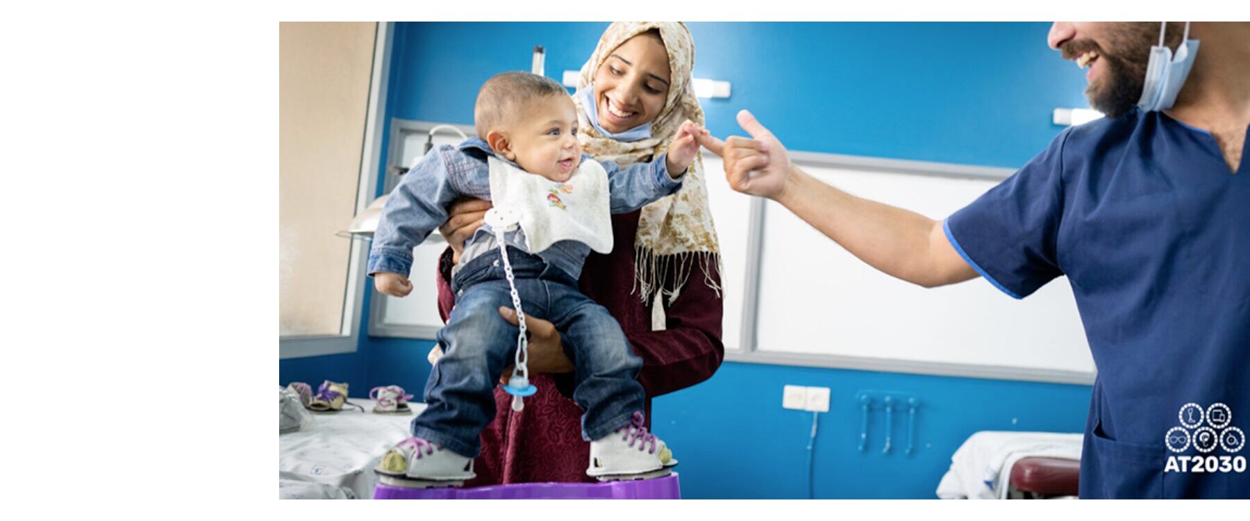 Mother holding child wearing a Ponseti brace between his feet. A medical person holds out a finger and the baby is touching it. All have smiles on their faces.