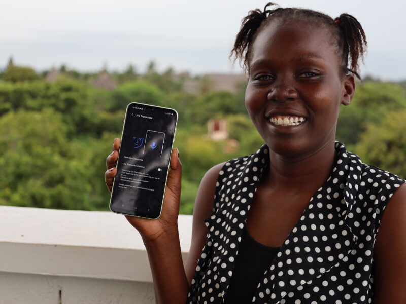 A lady holding a smartphone displaying the "Live Transcribe" app interface. She is wearing a black and white polka dot top and is standing on a balcony with lush green trees and a clear sky in the background.