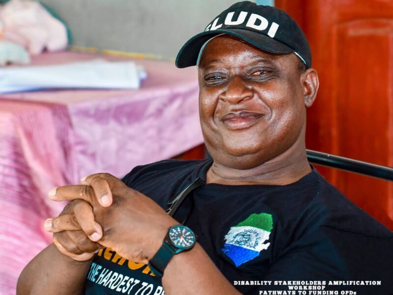 OPD leader in Sierra Leone - a man with a black cap smiling to camera while sitting indoors