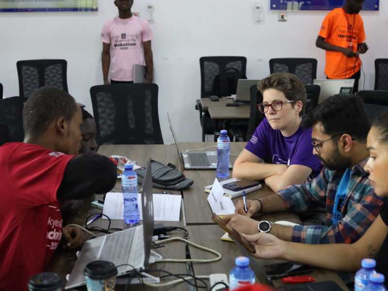 A group of people are seated at a table, working on laptops and papers. Some individuals are visible in the background, standing or seated at other tables.