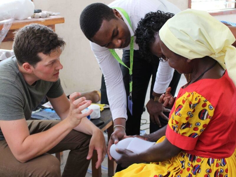 A group of four people, two men and two women, are gathered around a table. A man in a green t-shirt is sitting on the left, gesturing with his hands as he speaks to the others. In the center, a man in a white shirt and a woman with dark hair are leaning over the table, looking at an object. On the right, a woman in a red and yellow dress and a yellow headscarf is holding a white, curved object in her hands. She is seated, while the others are standing or crouching. The setting appears to be a workshop or studio, with various objects and tools visible on shelves and tables in the background.