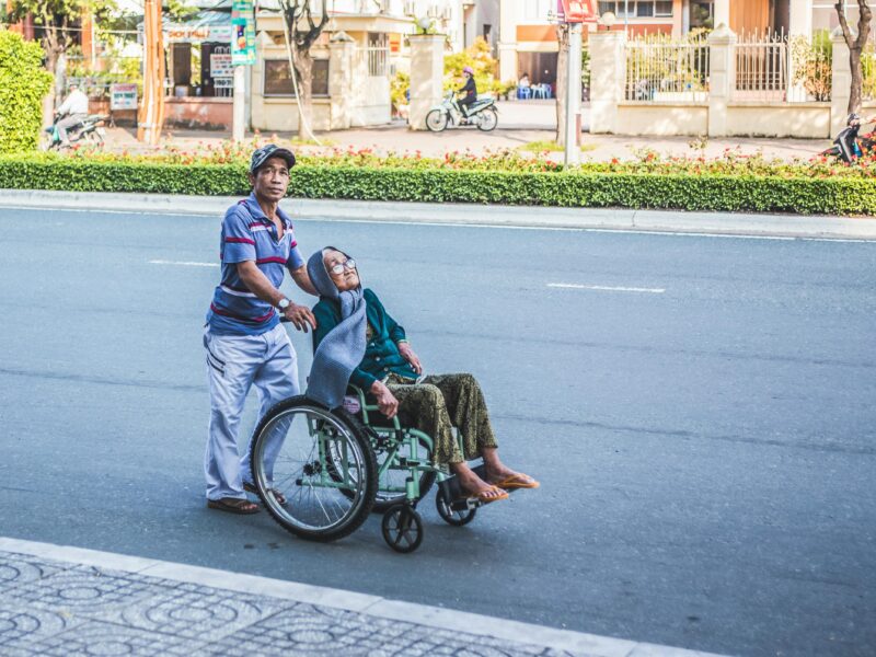 A person pushes another person in a wheelchair across a road, with buildings and trees in the background.