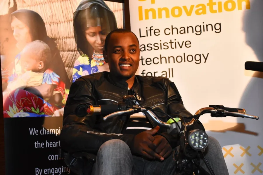 Lincoln, a young black man, wearing a leather jacket and sat astride a motorcycle at an event. Poster stands are behind him - one partially reads "Innovation. Life changing assistive technology for all"