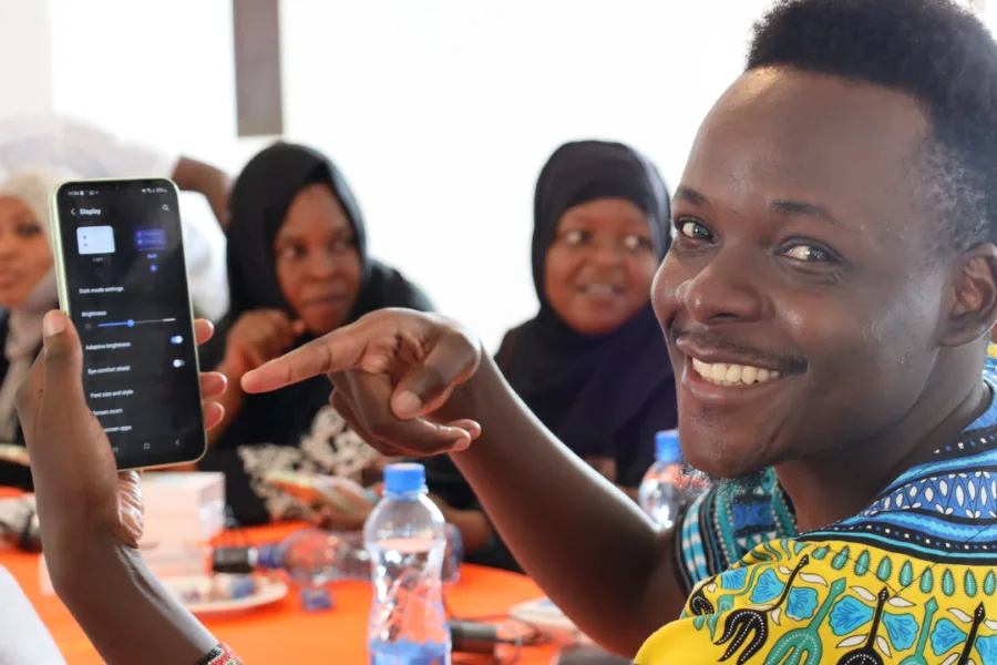 Image in Kenya of a trainer showcasing accessible features on a mobile. Male training is smiling warmly to the camera, in a bright printed top - and pointing to mobile phone.