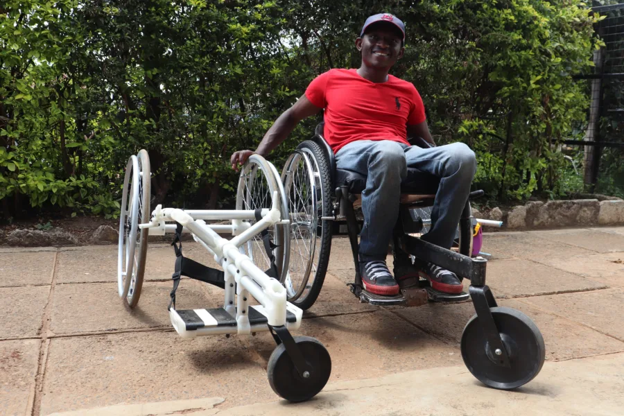 A smiling man wearing a red shirt and cap sits outdoors in a custom-built wheelchair with extra large wheels, designed for mobility on rough terrain. Greenery forms the background.