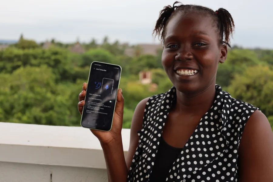 A lady holding a smartphone displaying the "Live Transcribe" app interface. She is wearing a black and white polka dot top and is standing on a balcony with lush green trees and a clear sky in the background.