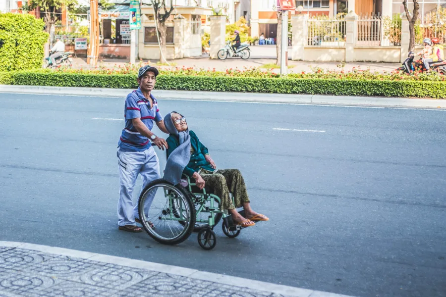 A person pushes another person in a wheelchair across a road, with buildings and trees in the background.