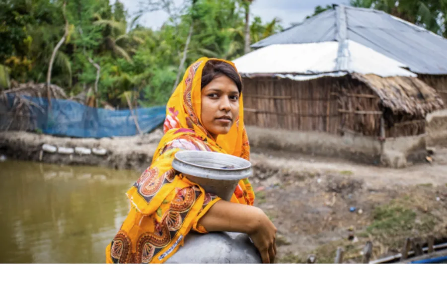 Person in a colorful garment stands near water with a rural house in the background; cover of the "Advancing Disability Inclusive Climate Action" guide featuring logos of CBM Global, IAG, and GDI Hub.