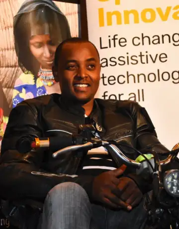 Lincoln, a young black man, wearing a leather jacket and sat astride a motorcycle at an event. Poster stands are behind him - one partially reads "Innovation. Life changing assistive technology for all"