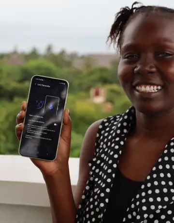 A lady holding a smartphone displaying the "Live Transcribe" app interface. She is wearing a black and white polka dot top and is standing on a balcony with lush green trees and a clear sky in the background.