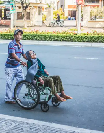 A person pushes another person in a wheelchair across a road, with buildings and trees in the background.