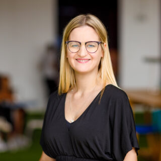 A white lady with brown hair , wearing glasses and a black top smiling at the camera