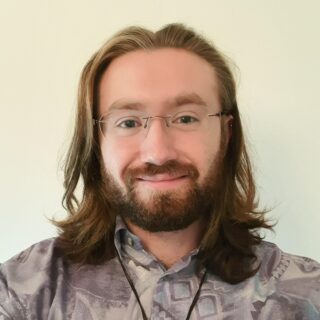 A photograph of Zak: a young white male with shoulder length hair and a beard. Zak is wearing glasses.