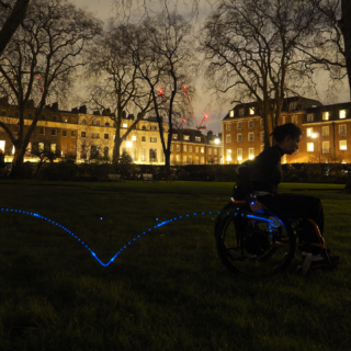 A view of a park in London at night. A manual wheelchair user is propelling a wheelchair in this park. The wheelchair user is wearing an inertial sensor in the wrist, a second one is attached to the wheel. A trail of blue light shows the trajectory that the sensor measures from a couple propulsions of the wheelchair.