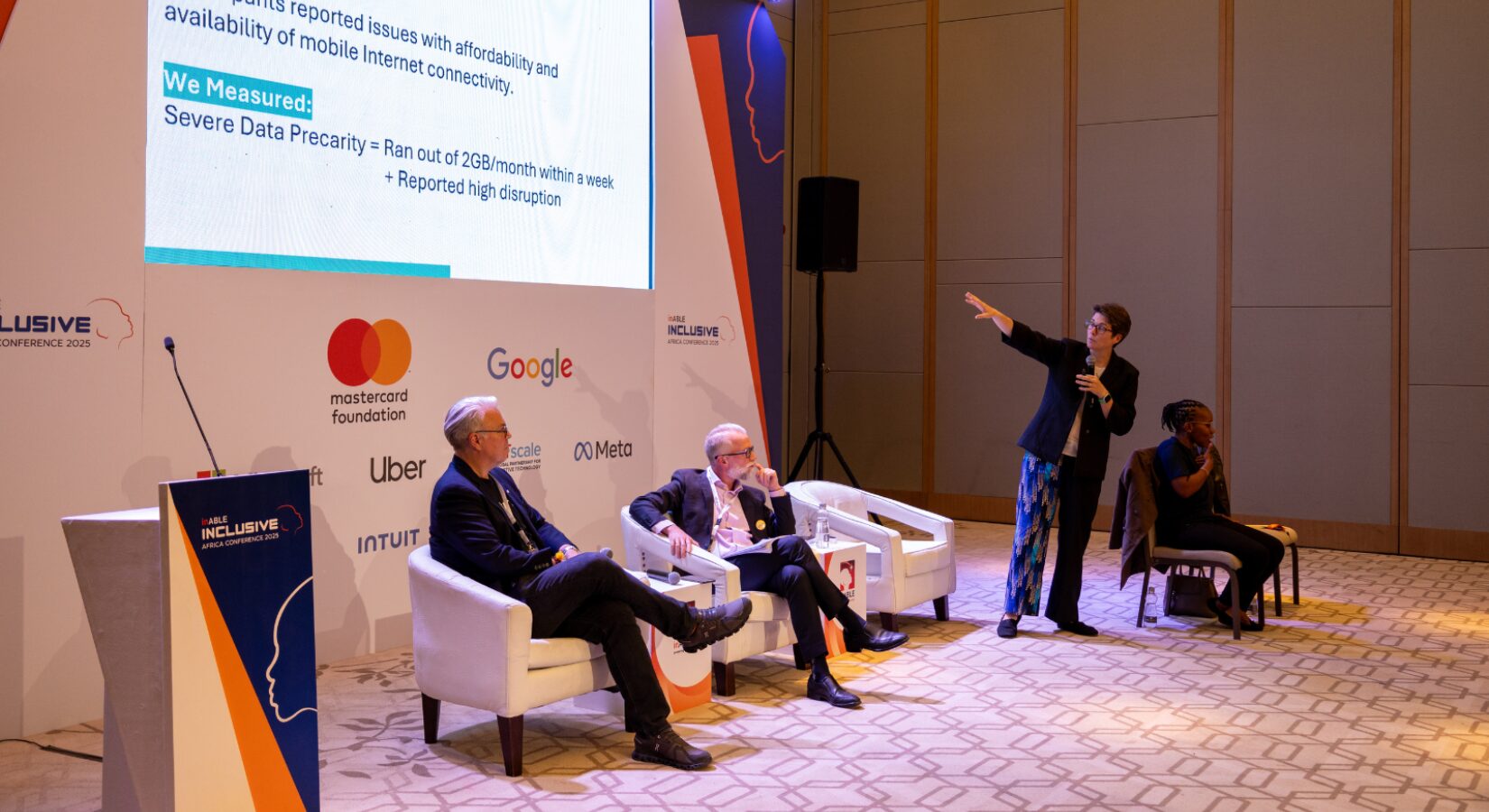 A panel discussion at a conference, with four people on a stage. One person is standing and speaking, while three others are seated. A large screen behind them displays a slide with text and logos, including Mastercard Foundation, Google, Uber, and Meta.