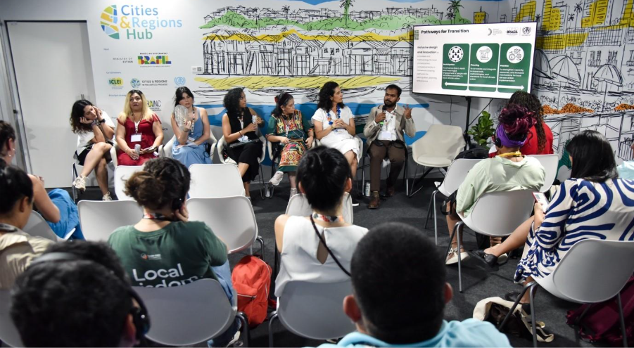 Panel discussion at the Cities & Regions Hub, COP-30, with speakers seated in a semicircle addressing an audience while a slide on “pathways for transition” displays three green diagrams.