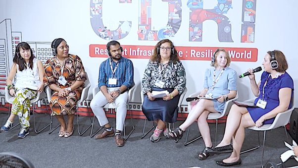 Six panelists sit in a row on white chairs; a woman on the far right speaks into a microphone while others listen with headsets and hold notes. A large backdrop reads “CDRI — Resilient Infrastructure: Resilient World,” with stylized city graphics.