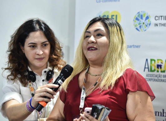 Two women at a conference booth, one holding a microphone toward the other, with event logos on the backdrop behind them.
